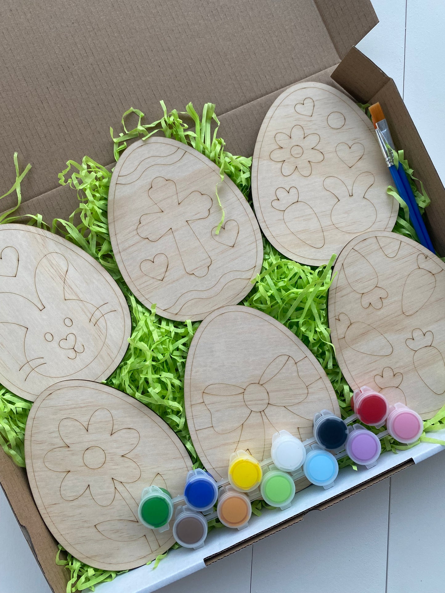 Wooden Easter egg-shaped crafts with paint supplies on a bed of green shredded paper.