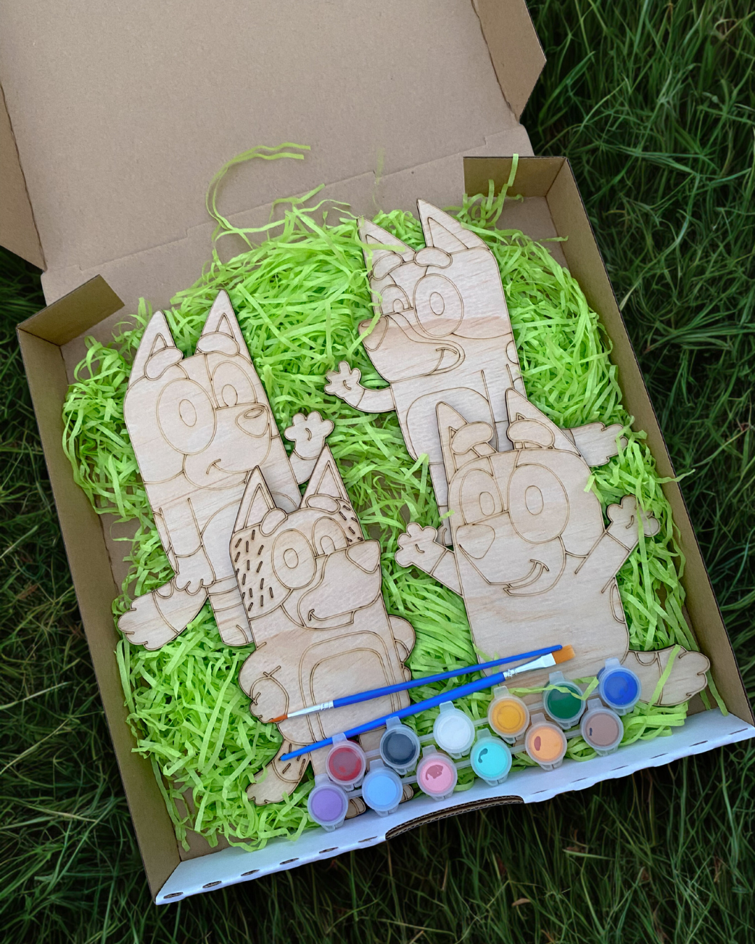 Wooden craft figures with paint supplies in a box on grass