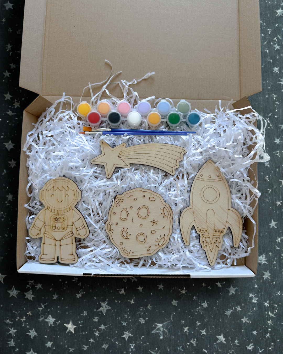 Box with wooden space-themed decorations including a astronaut, moon, and rocket ship, along with paint and brushes.