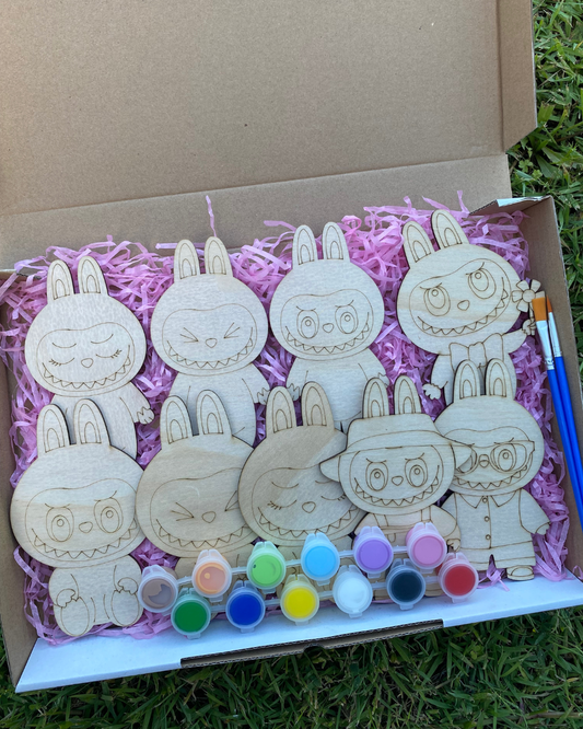 Set of wooden Labubu shapes with paint bottles and a brush in a box on grass