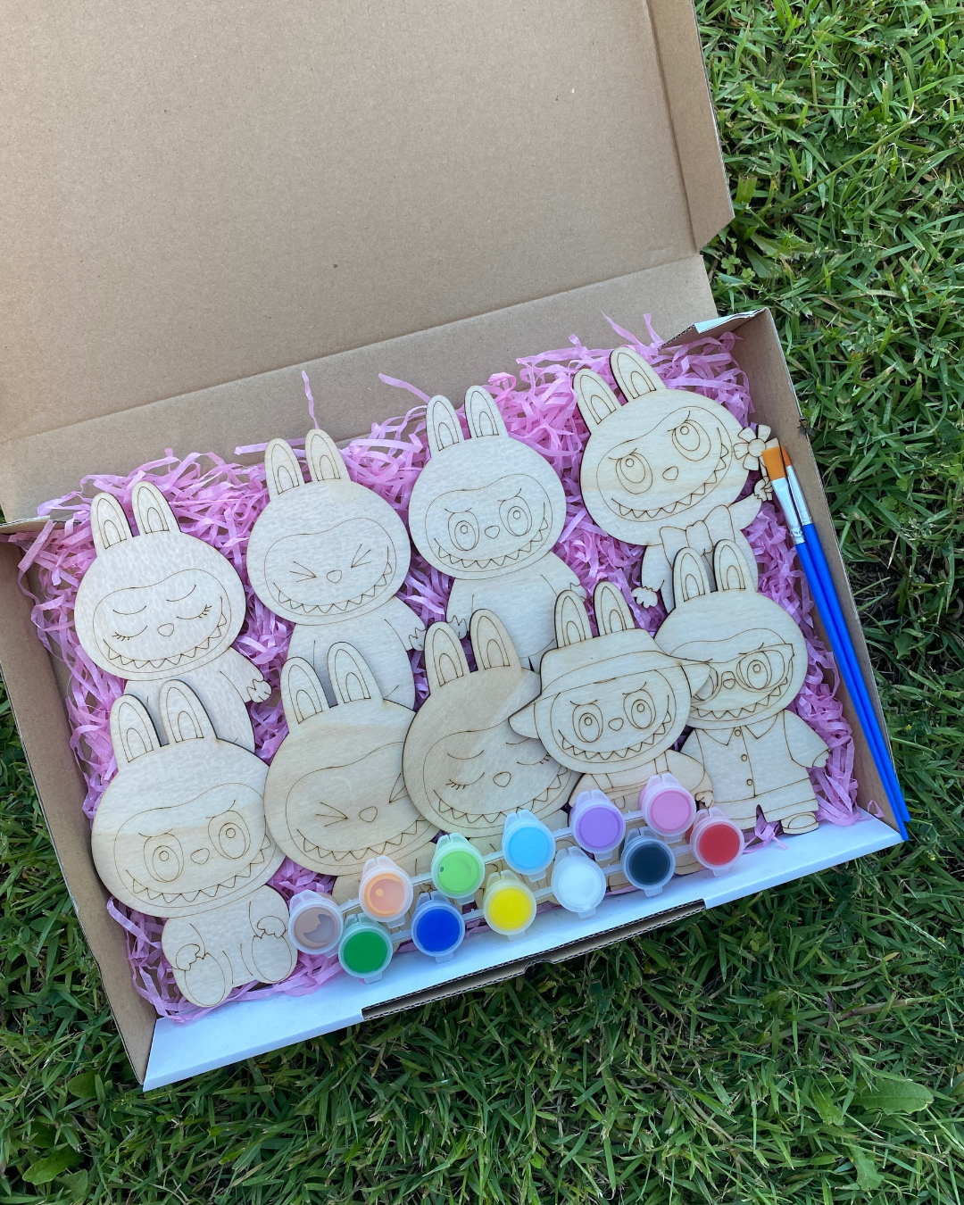 Box of wooden Labubu shapes with paint supplies on a grassy background
