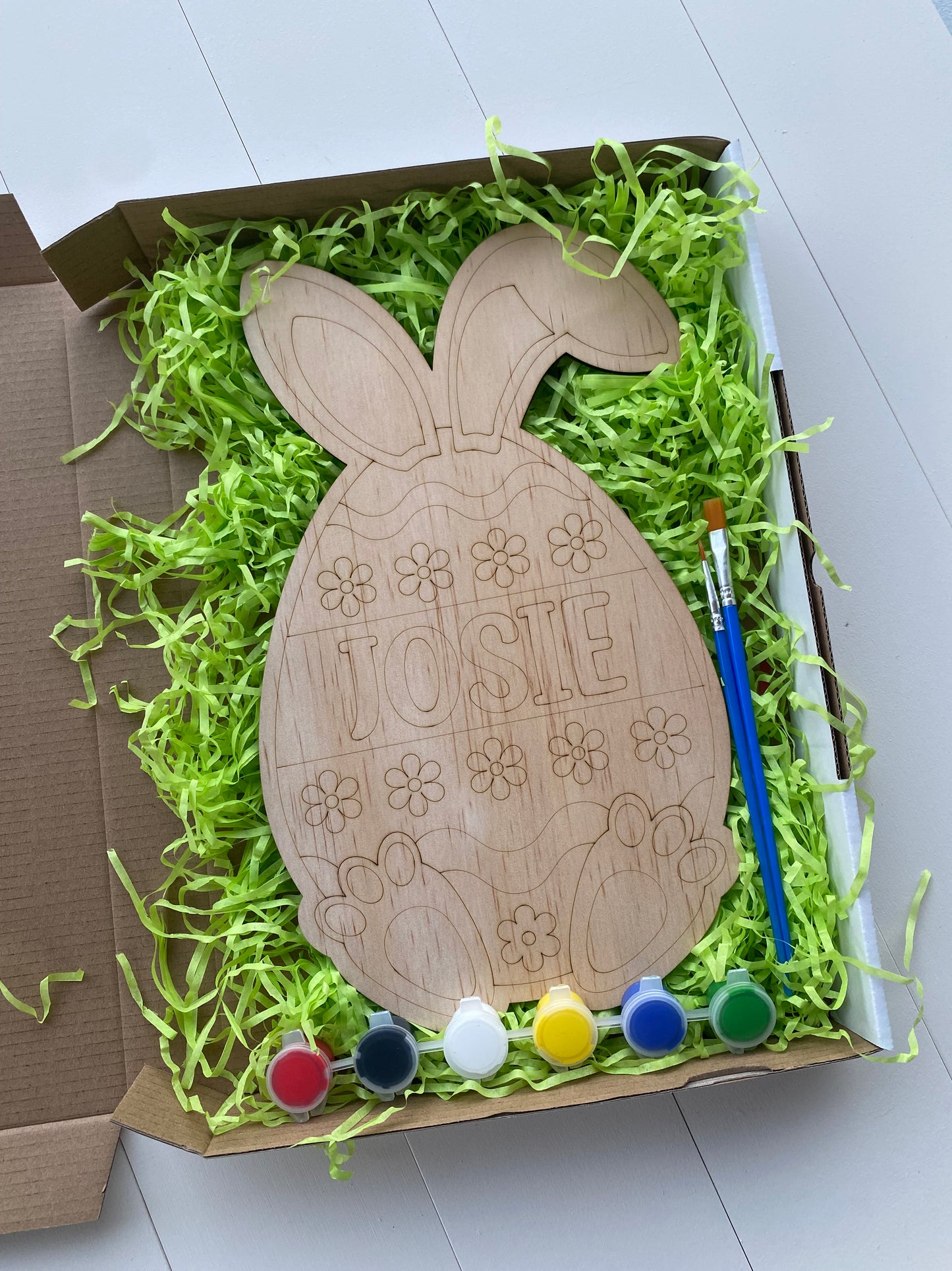 Wooden Bunny-shaped ornament with 'Josie' Etched onto the middle, inside a box filled with green shredded paper and paint supplies.