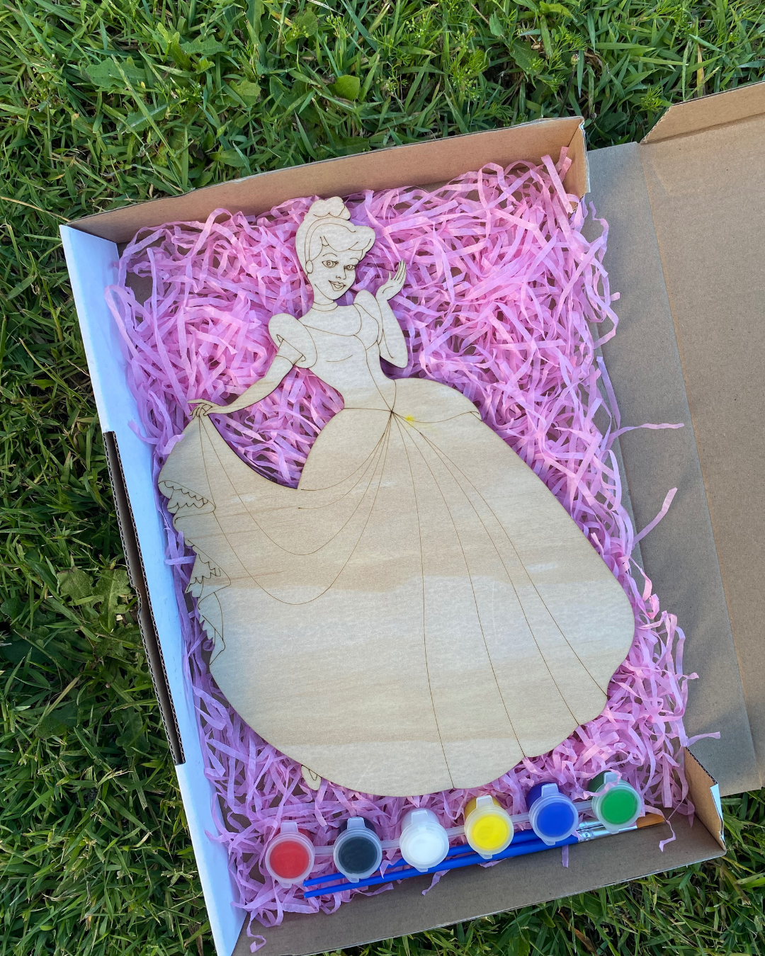 Wooden Cinderella figure in a box with pink filler and paint supplies on grass