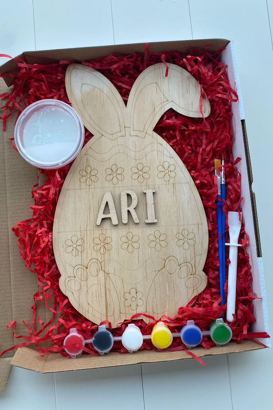 Wooden Bunny-shaped ornament with 'ARI' on a box filled with red shredded paper and paint supplies.