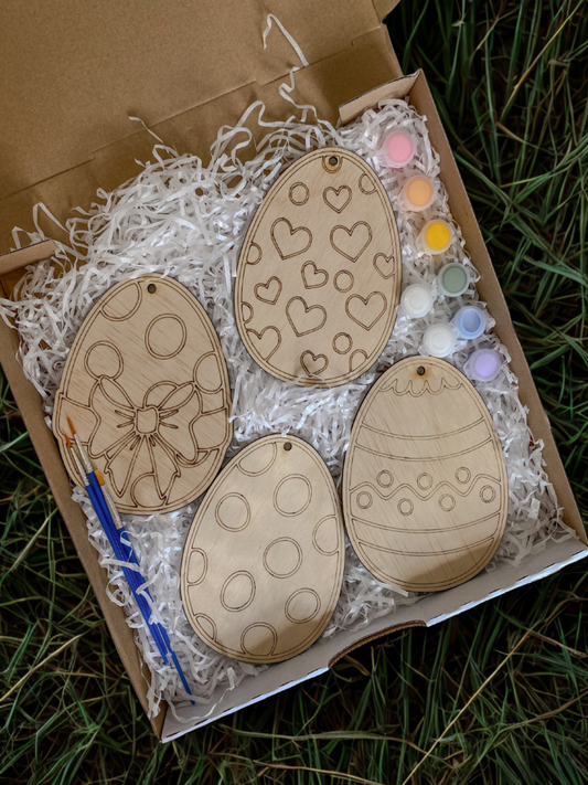 Wooden Easter egg ornaments with patterns in a box with paint supplies on grass.