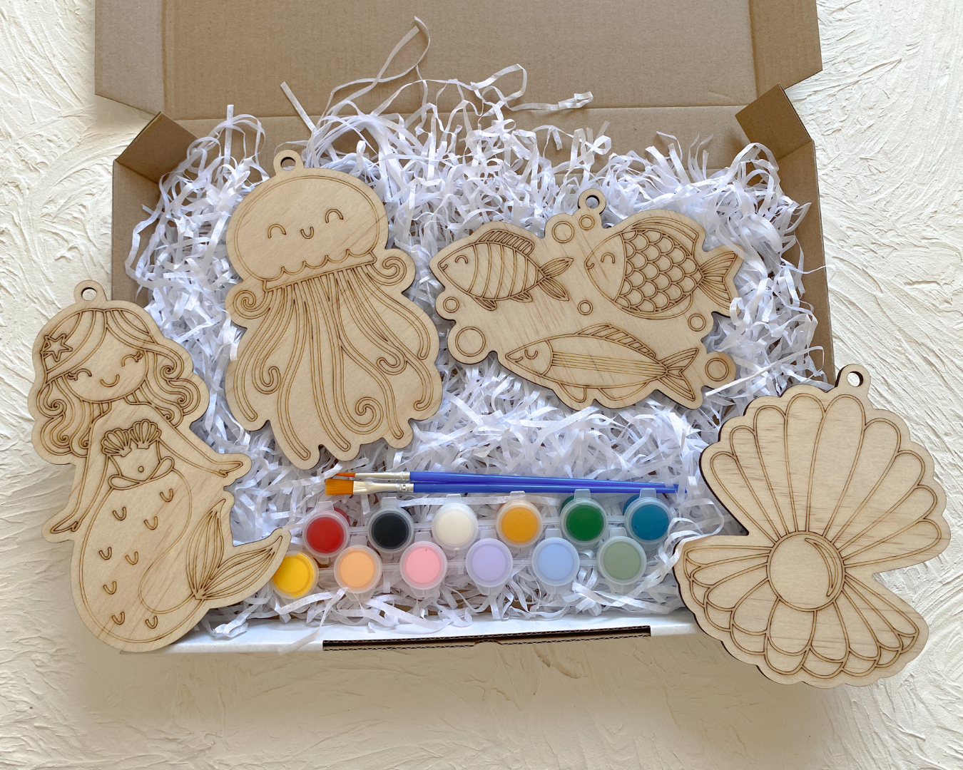 Wooden sea-themed craft set with paint, brushes, and wooden shapes on white shredded paper.