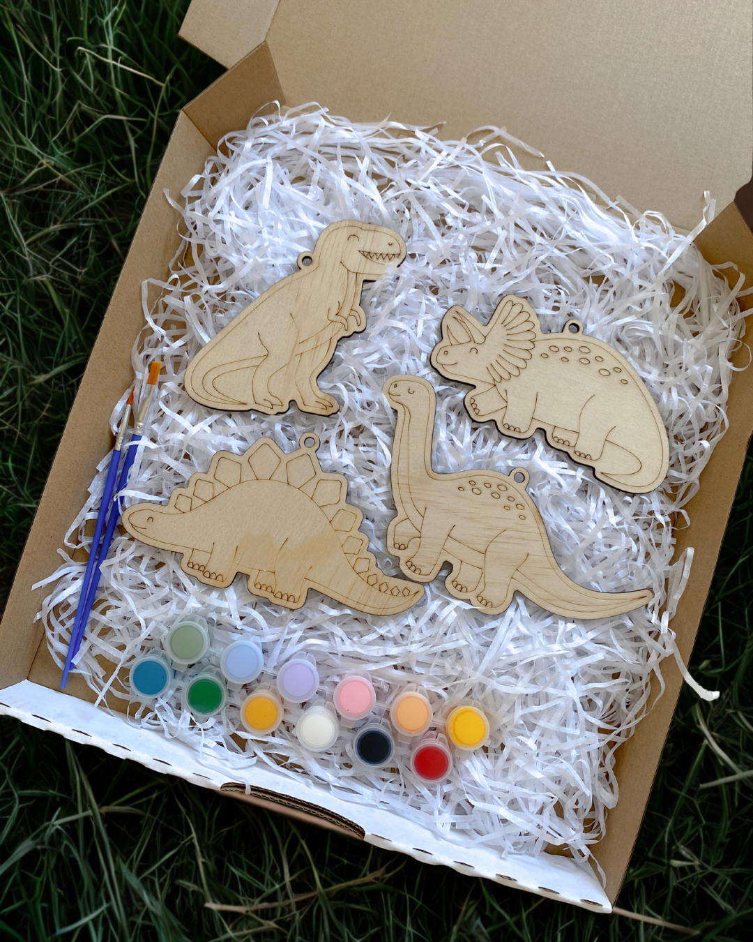 Wooden dinosaur shapes with paint supplies in a box on grass
