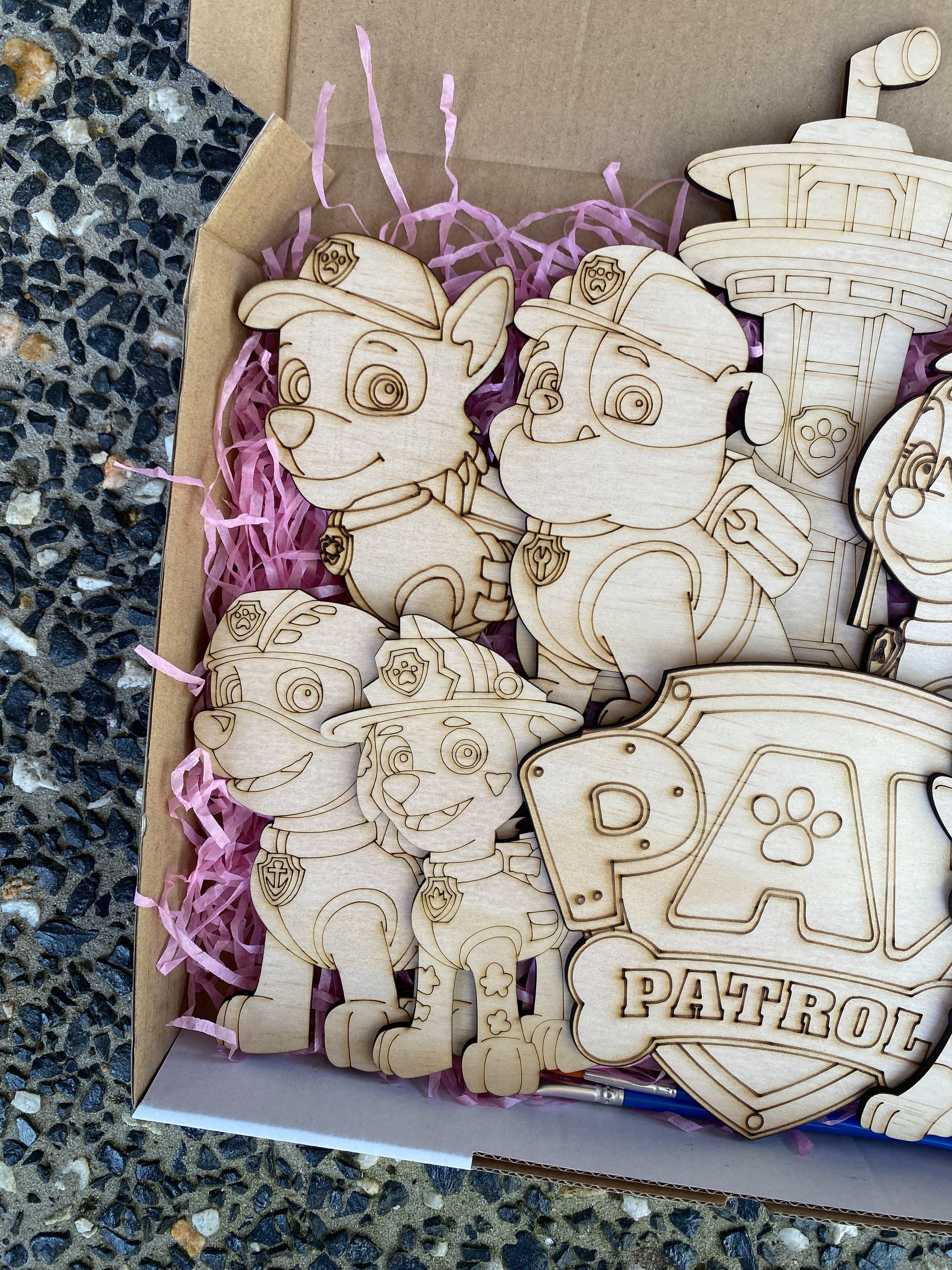 Wooden toys resembling characters from a popular children's show, displayed in a box with purple tissue paper.