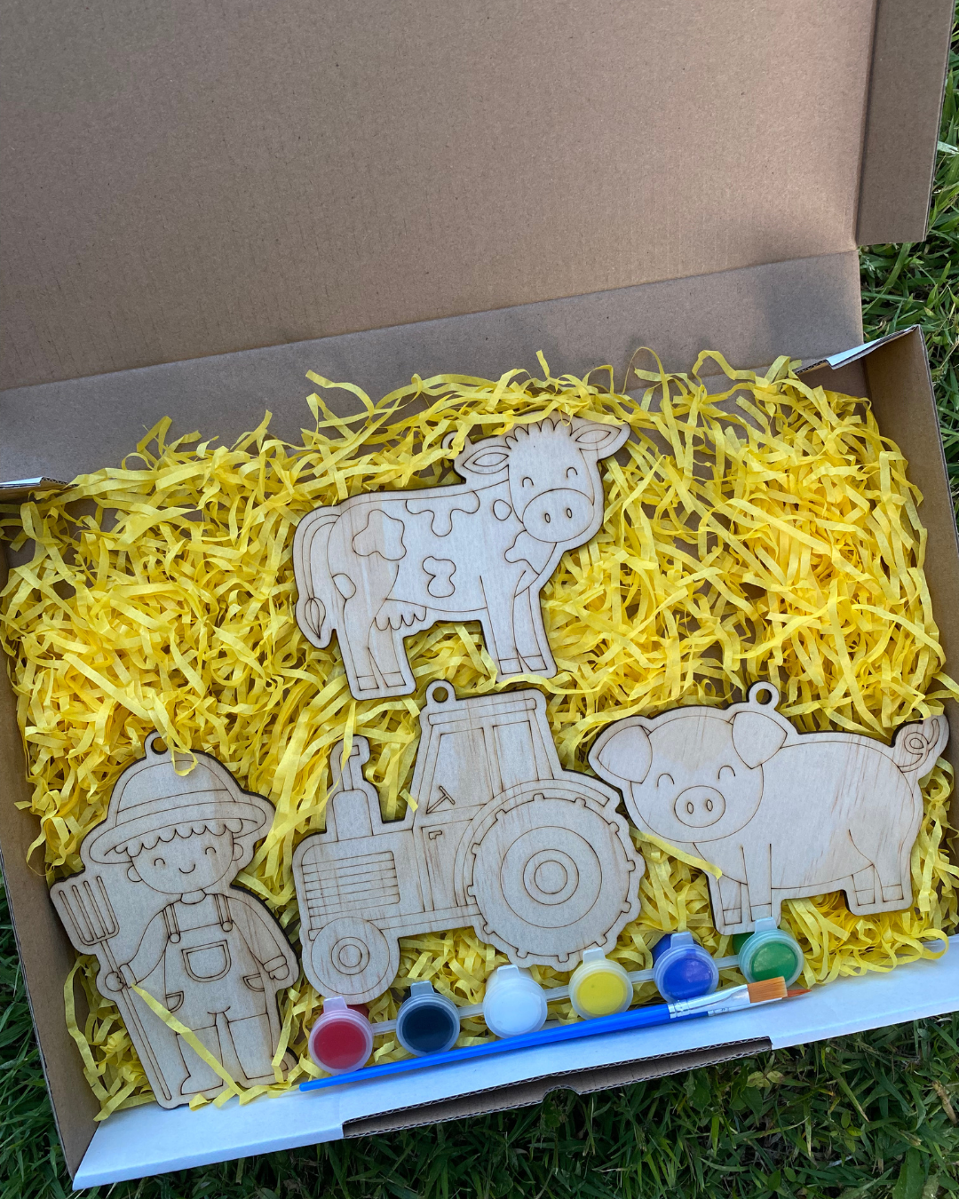 Wooden farm animal toys with paint supplies in a box filled with yellow shredded paper.