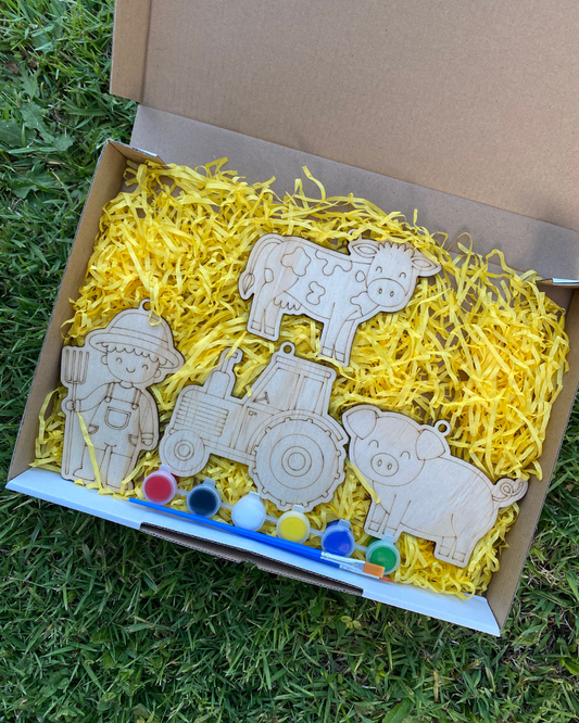 Wooden farm animal toys in a box with yellow filler and paint supplies on grass.
