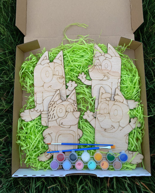 Wooden animal figures with paint supplies in a box filled with green shredded paper.