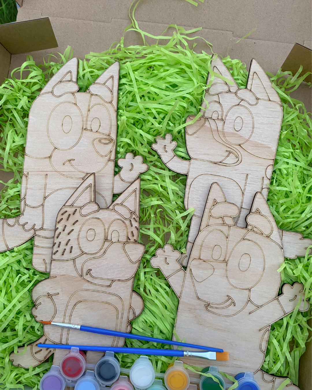 Wooden cutouts of cartoon characters with paint supplies on green shredded paper